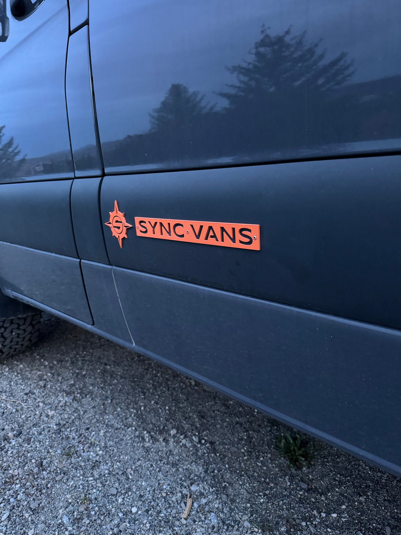 Dark van featuring an orange "SYNC VANS" logo, suggesting a custom RV rental, parked on gravel for outdoor adventures.