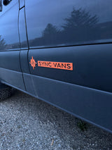 Dark van featuring an orange "SYNC VANS" logo, suggesting a custom RV rental, parked on gravel for outdoor adventures.