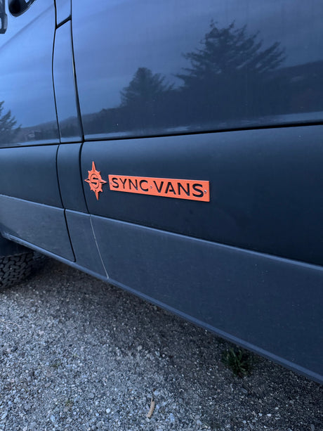 Dark van featuring an orange "SYNC VANS" logo, suggesting a custom RV rental, parked on gravel for outdoor adventures.