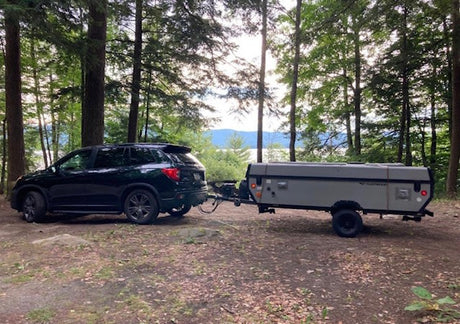 A dark SUV is hitched to an rv rental pop-up camper trailer, parked in a wooded area with a lake in the background.
