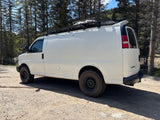 White camper van, equipped with a roof rack and off-road tires, parked on a forest dirt road. Ideal for an RV rental adventure.