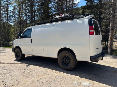 White camper van, equipped with a roof rack and off-road tires, parked on a forest dirt road. Ideal for an RV rental adventure.
