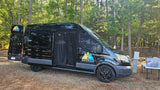 Black Wandervans camper van for rv rental, parked at a sunny wooded campsite with an open side door.