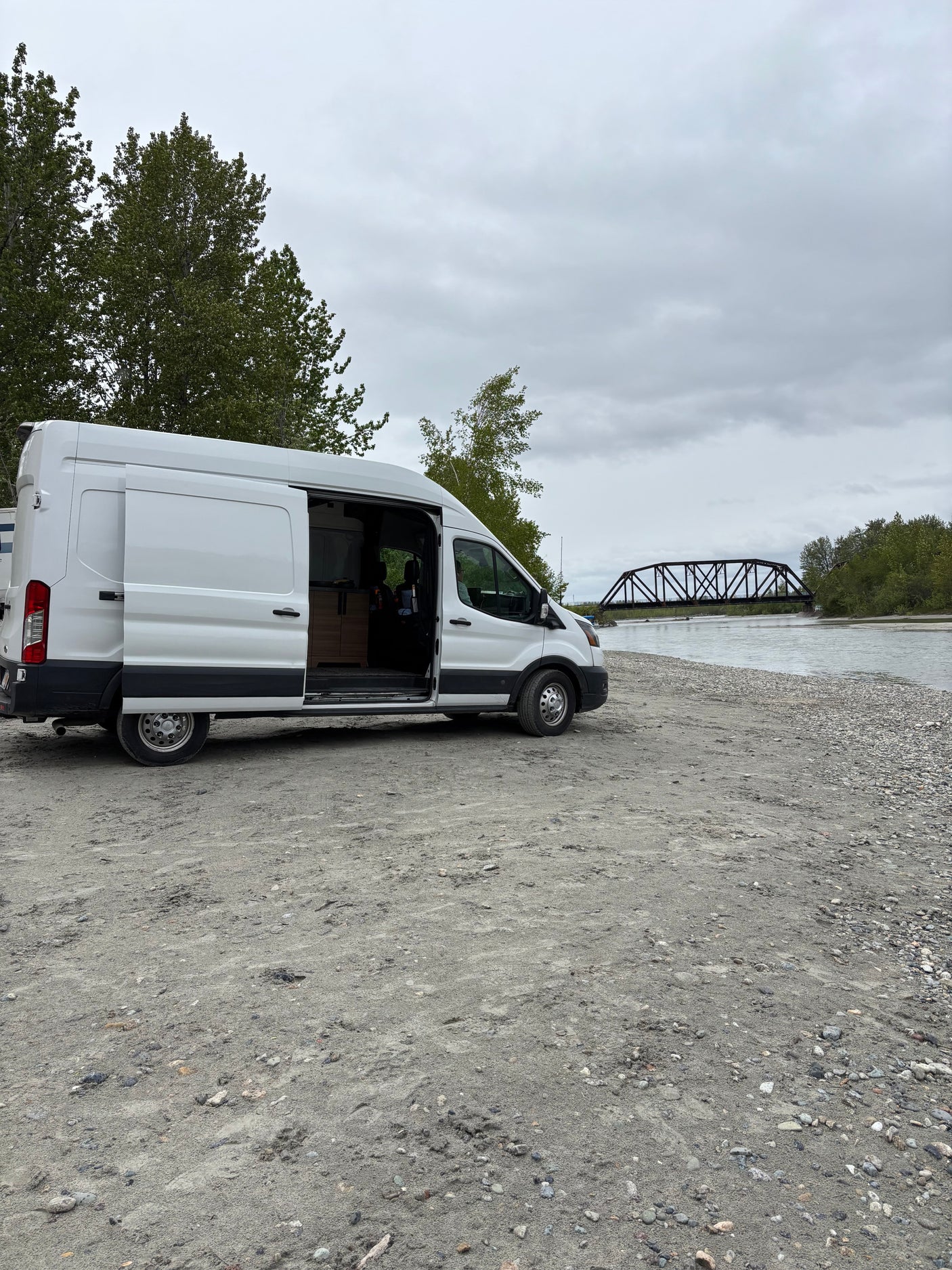 Riverside RV rental: A white camper van with its door open, parked on a gravel bank overlooking a river and bridge.