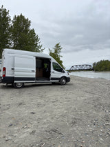 Riverside RV rental: A white camper van with its door open, parked on a gravel bank overlooking a river and bridge.