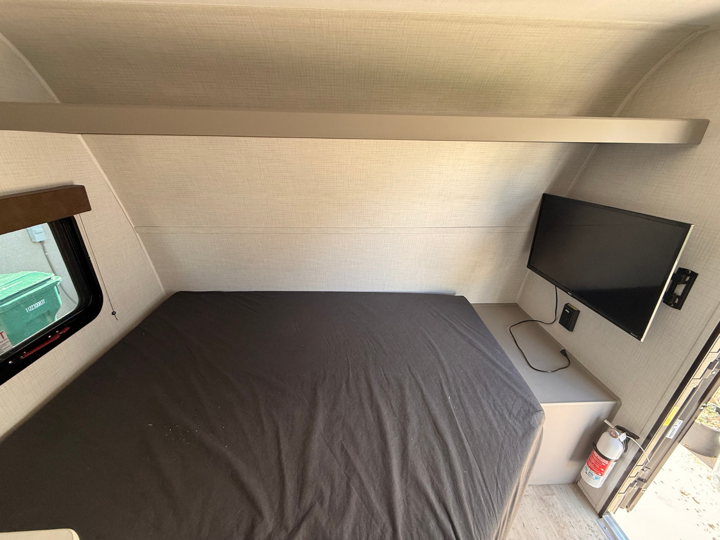 RV rental interior: cozy bedroom with dark bed, mounted TV, window showing outside, overhead shelf, and safety fire extinguisher. — photo 5
