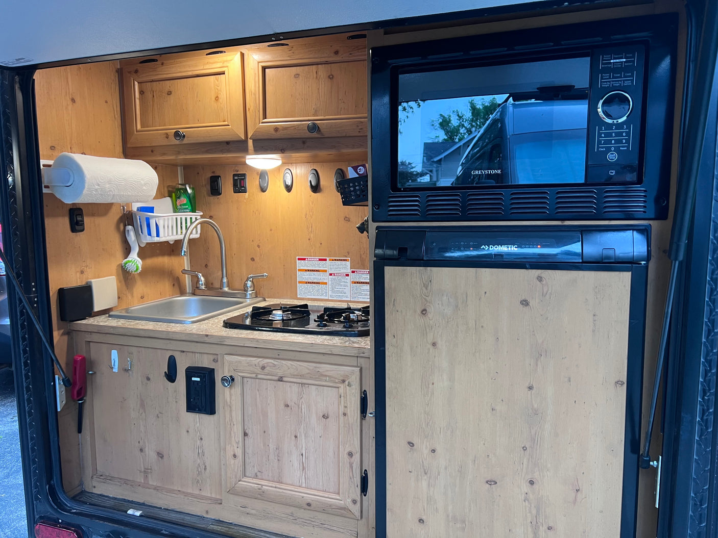Inside an RV rental: a compact camper kitchen with sink, stovetop, Greystone microwave, and Dometic fridge.