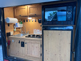 Inside an RV rental: a compact camper kitchen with sink, stovetop, Greystone microwave, and Dometic fridge.