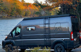 A dark grey camper van, featuring a Maine map graphic, parked by autumn-colored trees and water. Ideal for an RV rental getaway.