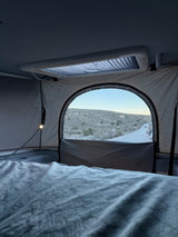 Inside an RV rental: a comfy bed, small lamp, and stunning desert view from the arched window. Ready for camping!