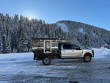 Silver truck with a black Four Wheel Campers pop-top camper, ready for an RV rental adventure, parked on an icy road in snowy mountains.