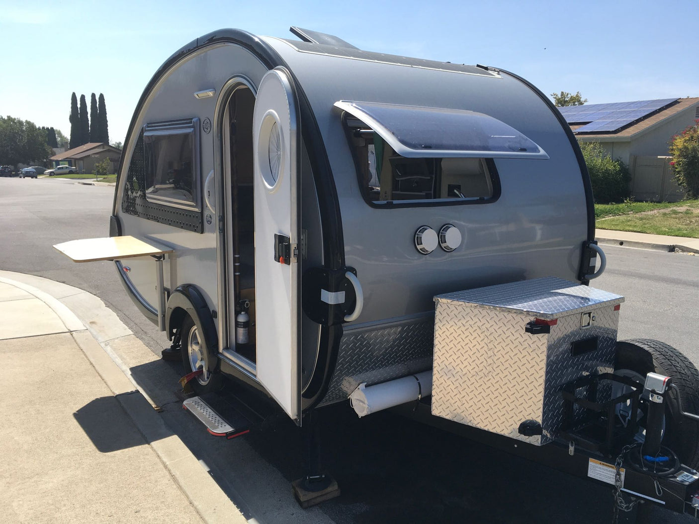 Compact grey teardrop RV rental parked on a sunny residential street with door open, showing interior and solar panels.