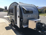 Compact grey teardrop RV rental parked on a sunny residential street with door open, showing interior and solar panels.