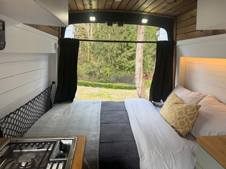 RV rental interior: cozy bed, compact kitchen stove, and serene forest view from open rear doors of this camper van.