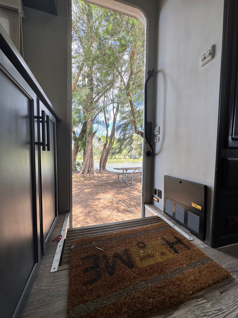 Serene rv rental camping view: an open door reveals trees, water, and a picnic table. A "HOME" mat is on the floor. — photo 17