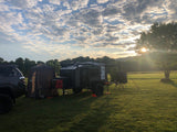 Teardrop rv rental & SUV parked at a grassy campsite. Beautiful sky with clouds and bright sun during golden hour.