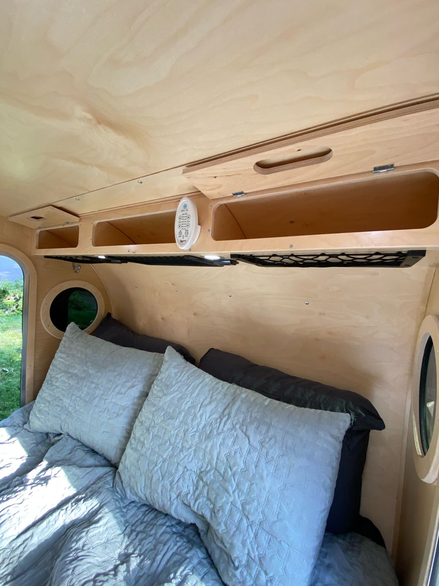 Compact RV rental bedroom with light wood paneling, cozy bed, overhead storage, and control panel.