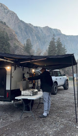 A person prepares a meal outside an RV rental, equipped with an awning and outdoor kitchen, at a mountain campsite.
