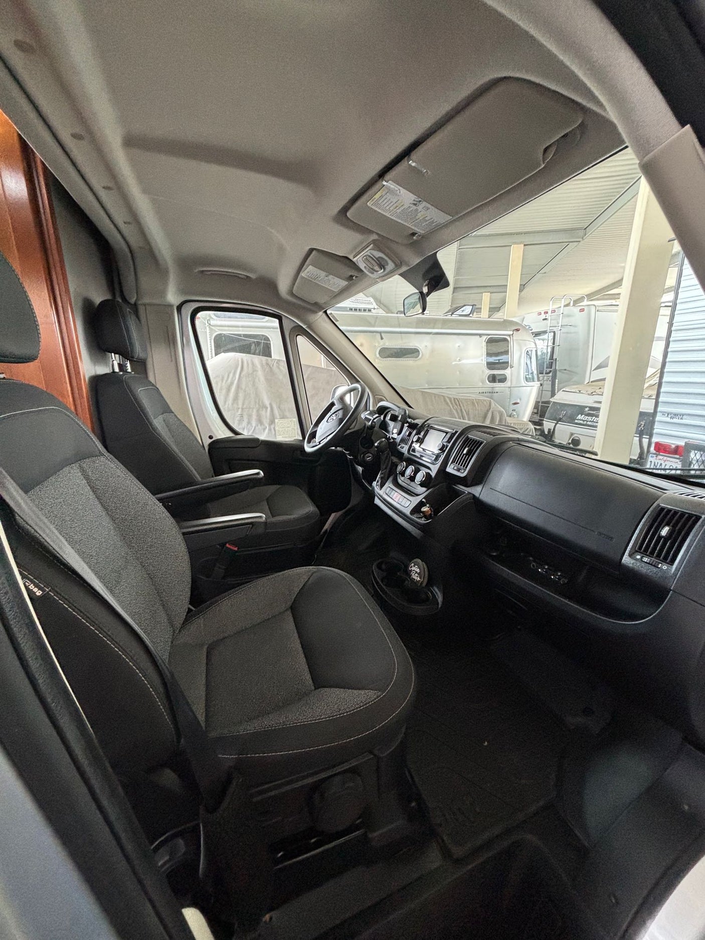 Inside an RV rental van: the front cabin with driver and passenger seats, and dashboard. Other RVs are visible through the windshield.