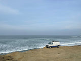 An RV rental, a white and blue van, is parked on a sandy cliff overlooking the ocean under cloudy skies.