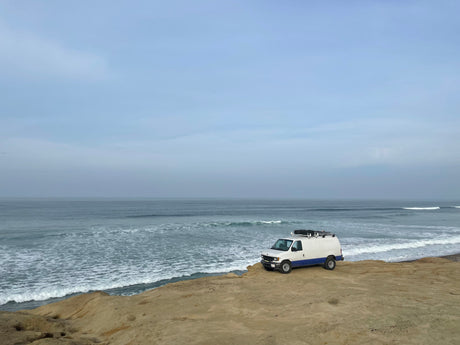 An RV rental, a white and blue van, is parked on a sandy cliff overlooking the ocean under cloudy skies.