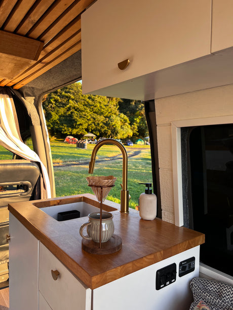 Bright RV rental interior with a wood-accented kitchen, brass faucet, pour-over coffee, and a view of a sunny campground.