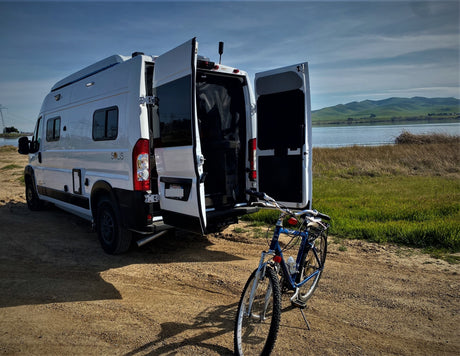 White RV rental van with open rear doors parked by a scenic lake. A blue bicycle stands ready for adventure.
