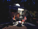 Enjoying an rv rental night: two people beside a bright campfire, camper van lights up the scene under dark trees.