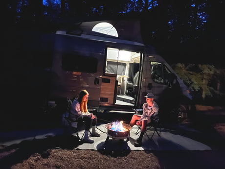 Enjoying an rv rental night: two people beside a bright campfire, camper van lights up the scene under dark trees.