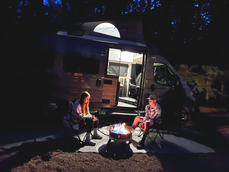 Enjoying an rv rental night: two people beside a bright campfire, camper van lights up the scene under dark trees. — photo 20