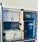 Interior of an rv rental kitchen, featuring a black microwave, refrigerator, stainless steel sink, and white storage cabinets.