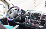 Inside an RV rental: driver's dashboard with a vibrant rainbow steering wheel cover, clear controls, and a hanging dreamcatcher.