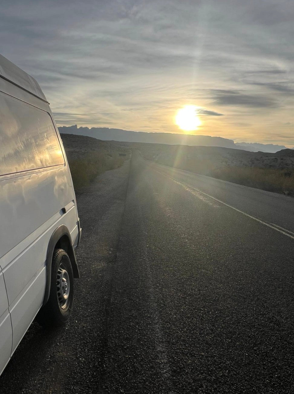 White RV rental van parked roadside, sun setting over distant mountains. Scenic road trip vibes.