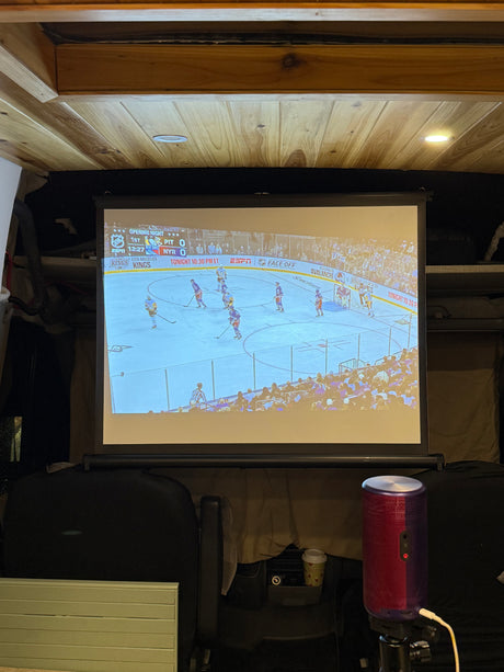 RV rental fun: A projector displays NHL hockey on a screen, offering cozy, on-the-go entertainment in a wood-paneled interior.