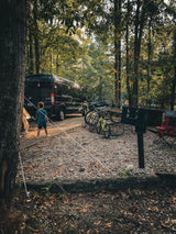 Family camping trip with an RV rental. A child stands near a camper van, bikes, and string lights at a wooded campsite.
