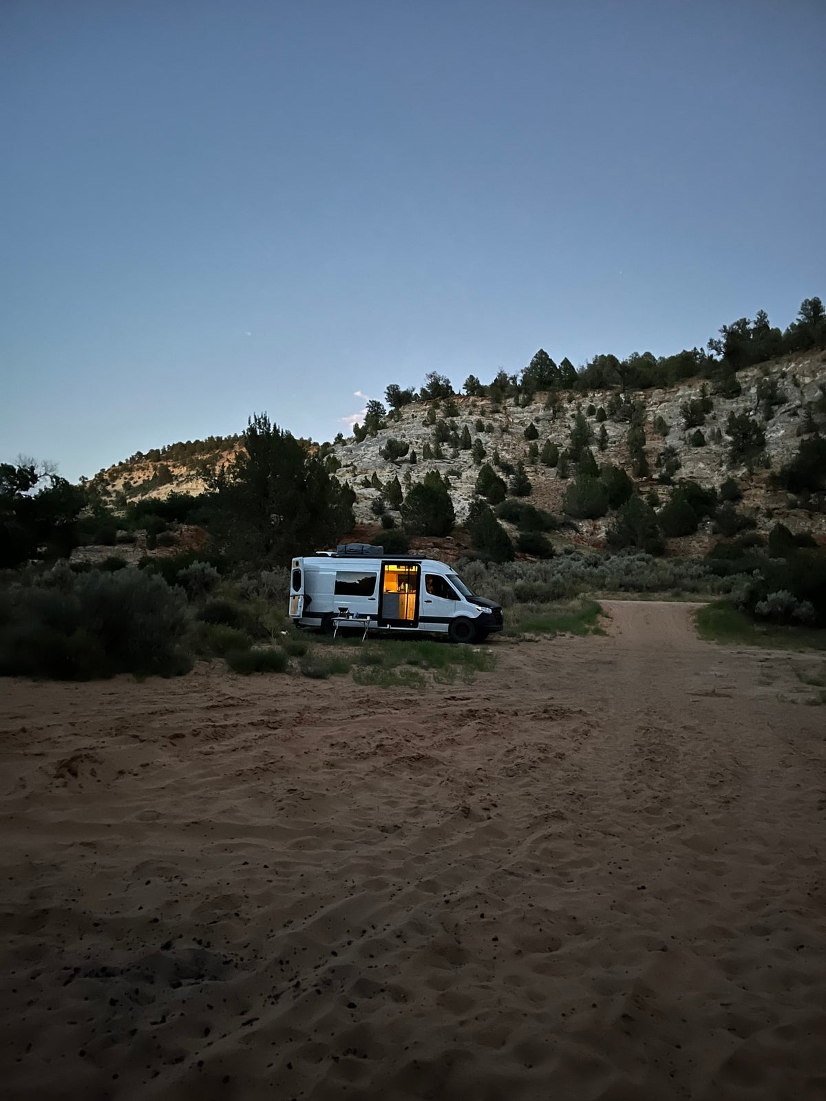 An RV rental camper van with interior lights on is parked at dusk on a sandy desert road near rocky hills, perfect for boondocking. — photo 18