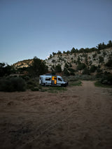 An RV rental camper van with interior lights on is parked at dusk on a sandy desert road near rocky hills, perfect for boondocking.