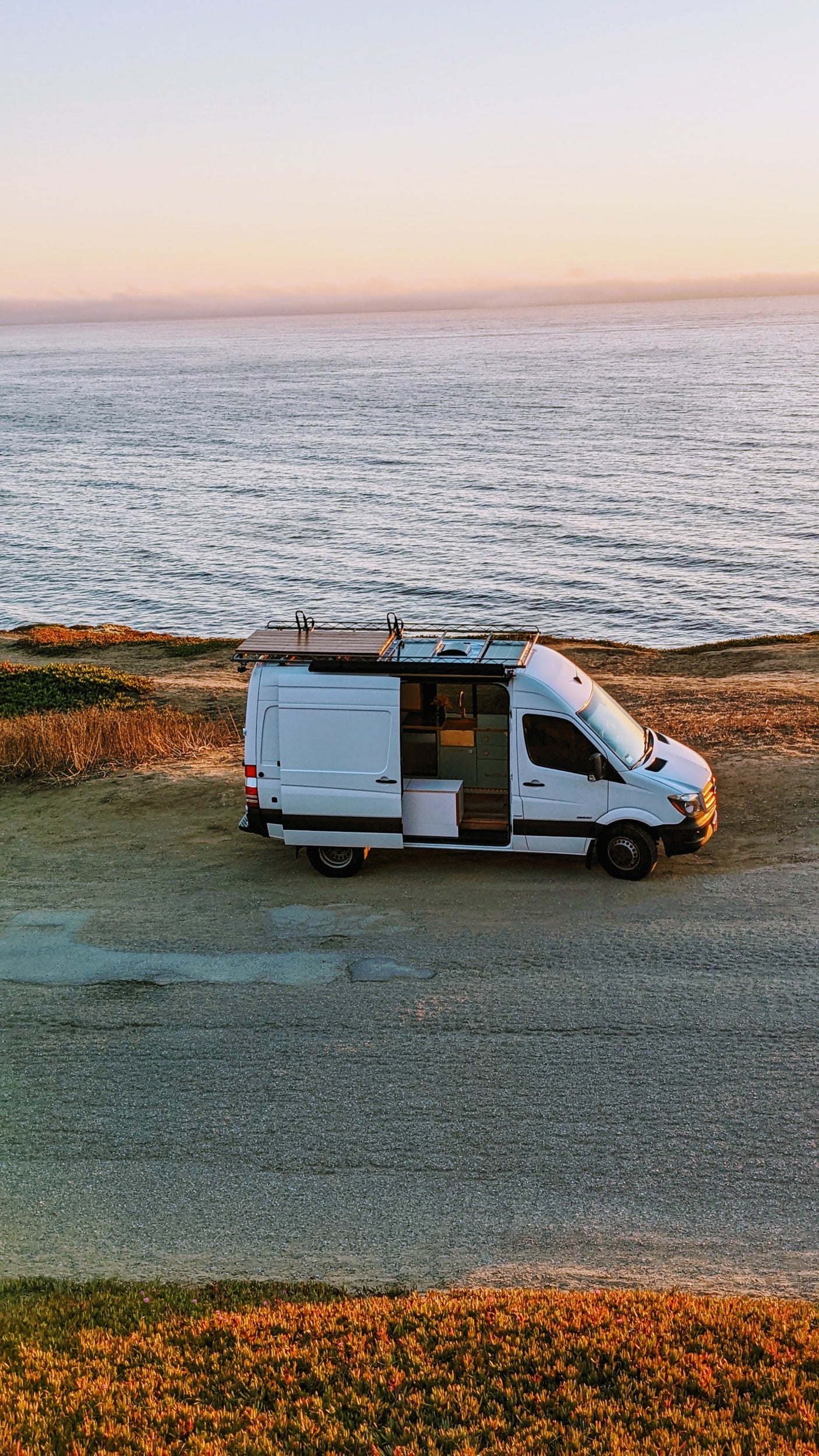White camper van with open door, revealing its interior, parked on a cliff above a calm ocean at sunset. Perfect for an RV rental.