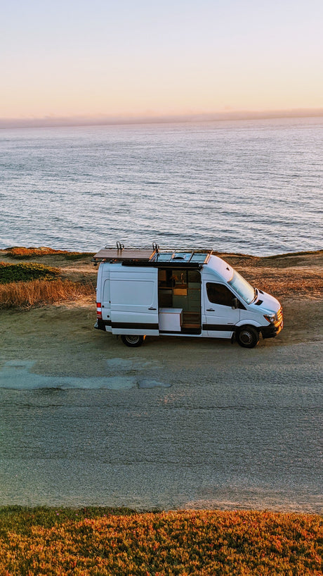 White camper van with open door, revealing its interior, parked on a cliff above a calm ocean at sunset. Perfect for an RV rental.