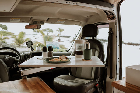 Inside an RV rental, a table is set with breakfast & coffee, overlooking a beautiful beach scene.
