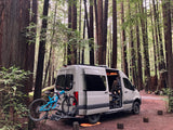 RV rental van parked in a serene redwood forest with bikes, ready for outdoor adventure and exploring nature.