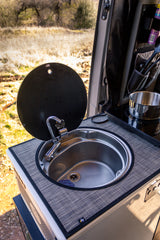 This RV rental's compact kitchen features a stainless steel sink with a faucet, a lift-up cover, and an induction cooktop with a pot, ready for meals in nature.