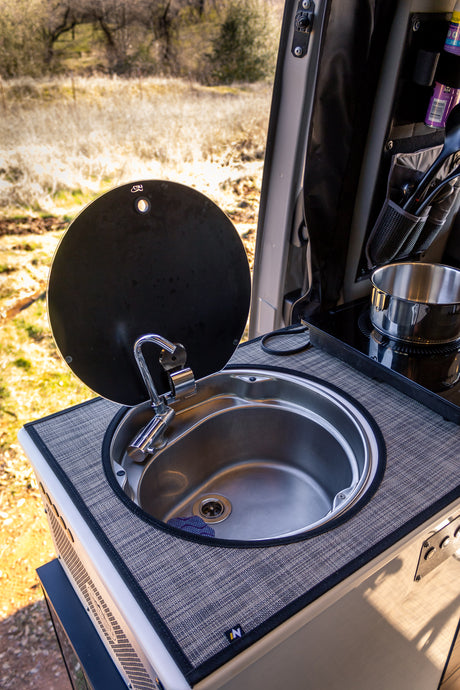 This RV rental's compact kitchen features a stainless steel sink with a faucet, a lift-up cover, and an induction cooktop with a pot, ready for meals in nature.
