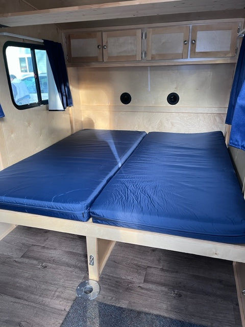 Inside a compact rv rental: two blue bunk beds on the right, light wood cabinets on the left, and a window below.