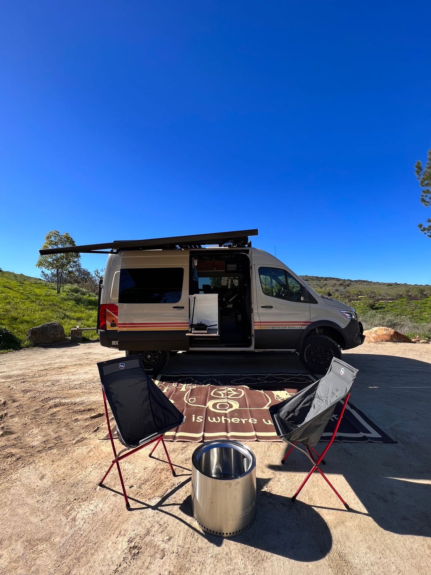 Perfect rv rental setup: camper van, chairs, Solo Stove fire pit on dirt with clear blue sky & green hills for an adventure.