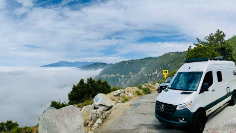 This rv rental offers breathtaking views! A white camper van overlooks mountains above a thick cloud layer from a scenic road.