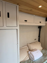 RV rental's cozy campervan interior with a bed, pillows, white towel, overhead storage, and wooden ceiling with a light.