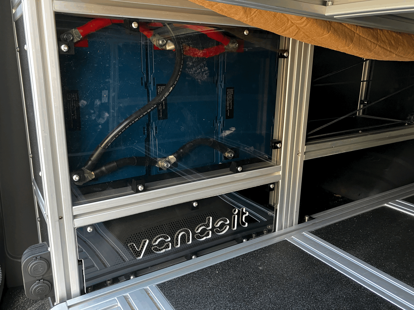 Inside an RV rental: a custom van's high-capacity lithium battery power system, visible through clear panel, with heavy-gauge cables.