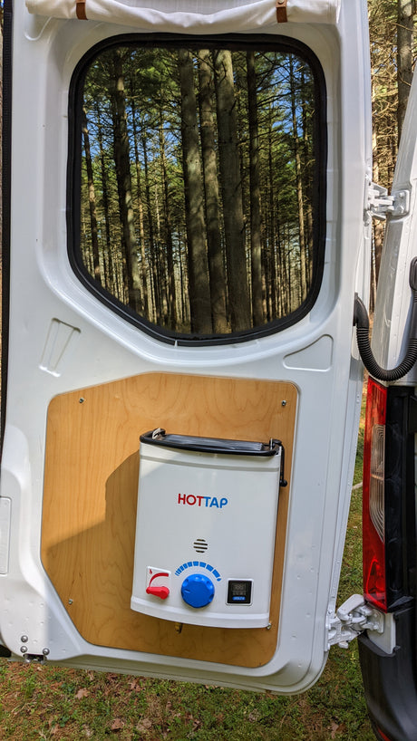 View from an open white **RV rental** van door, showing a mounted HOTTAP outdoor water heater. Through the window, a forest is visible.