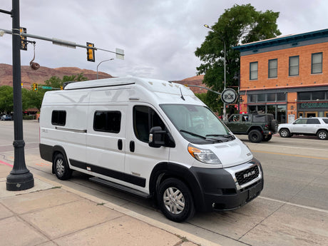 A white Winnebago Solis RV, often an RV rental, is parked on Center St. in Moab, Utah, with scenic mountains behind.